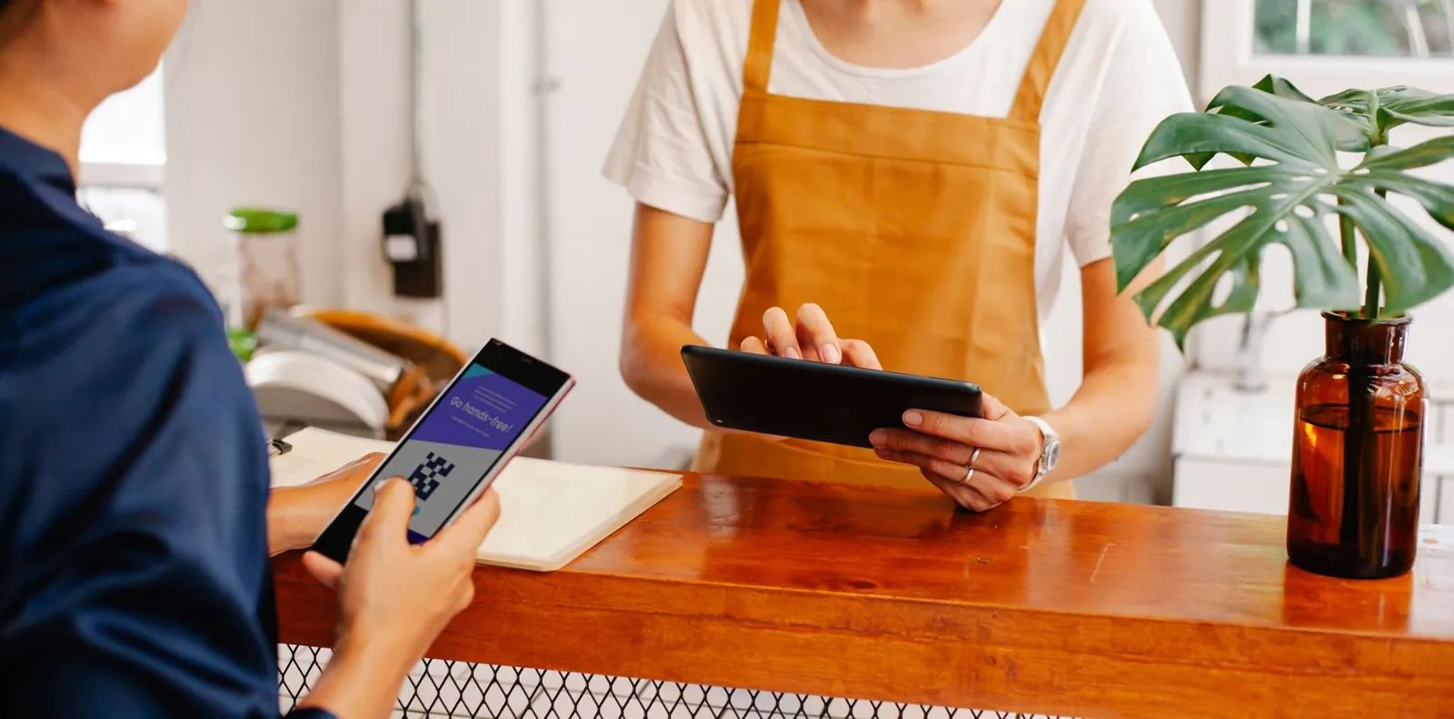 A customer holding a phone at a wooden cafe counter while staff use a tablet