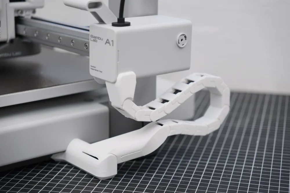 White articulated 3D-printed drag chain linking the side of a Bambu Lab A1 down to the bench, carrying the cable in a controlled loop