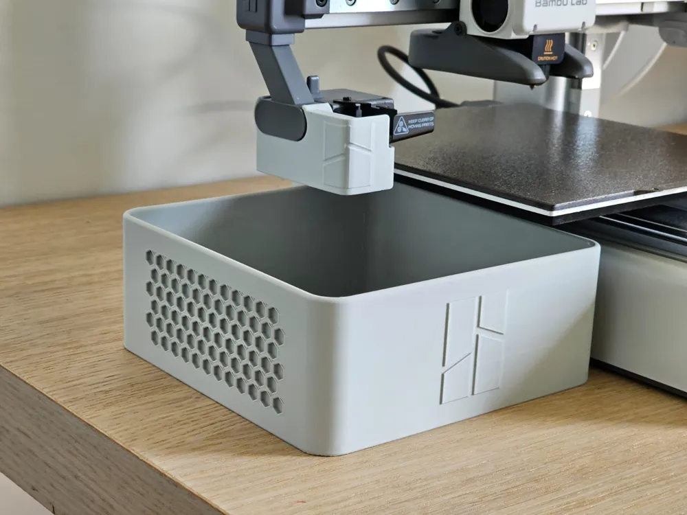 Grey 3D-printed poop bucket with a honeycomb vent panel sitting on a bench beside a Bambu Lab A1, with the deflector clipped to the toolhead above it