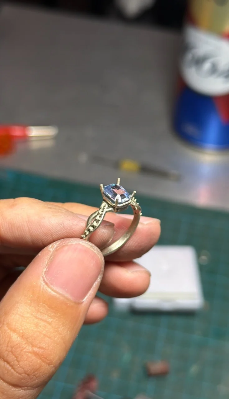 A finished ring with a light-coloured stone, held between fingertips at a workbench